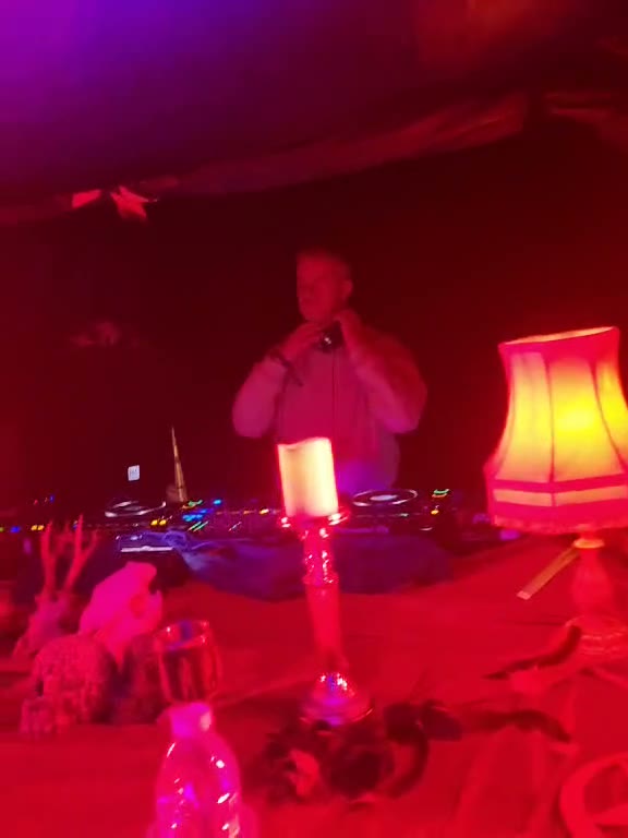 A DJ behind the decks in red light, a candle and a lamp lit on the table in front
